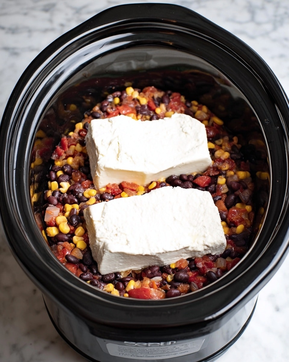A black slow cooker filled with a mix of black beans, corn, and diced tomatoes that create a colorful base layer. On top of this is a layer of two thick white cream cheese blocks, slightly soft and creamy in texture. The overall scene is set on a white marbled surface, with the slow cooker centered in the frame, showing a clear contrast between the dark cooker and bright ingredients inside. Photo taken with an iphone --ar 4:5 --v 7