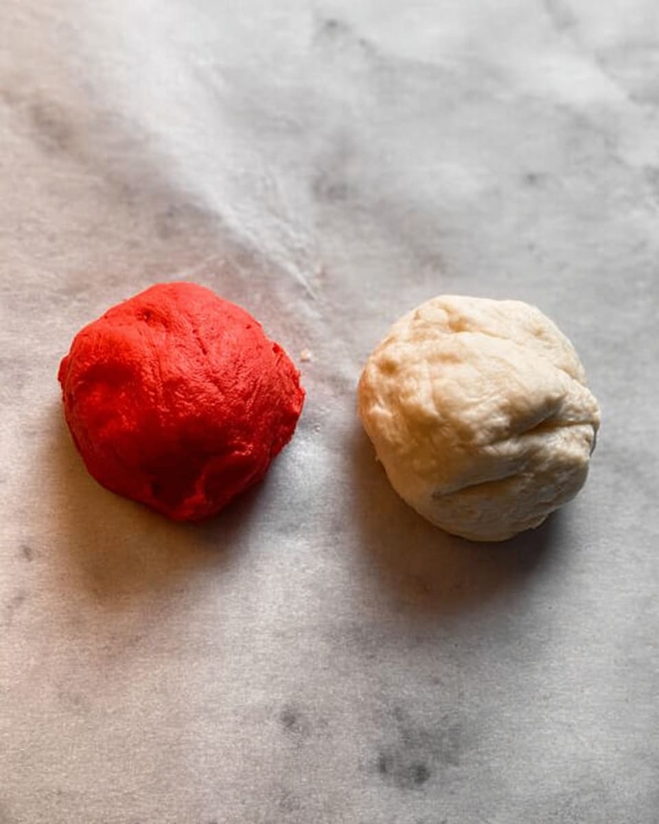 The image shows two small balls of dough placed side by side on a white marbled surface covered with parchment paper. The dough ball on the left is bright red, smooth but slightly uneven in texture with a few small bumps. The dough ball on the right is off-white with a soft, fluffy texture and a few creases on its surface. The balls are roughly the same size and positioned a little apart from each other on the paper. Photo taken with an iphone --ar 4:5 --v 7
