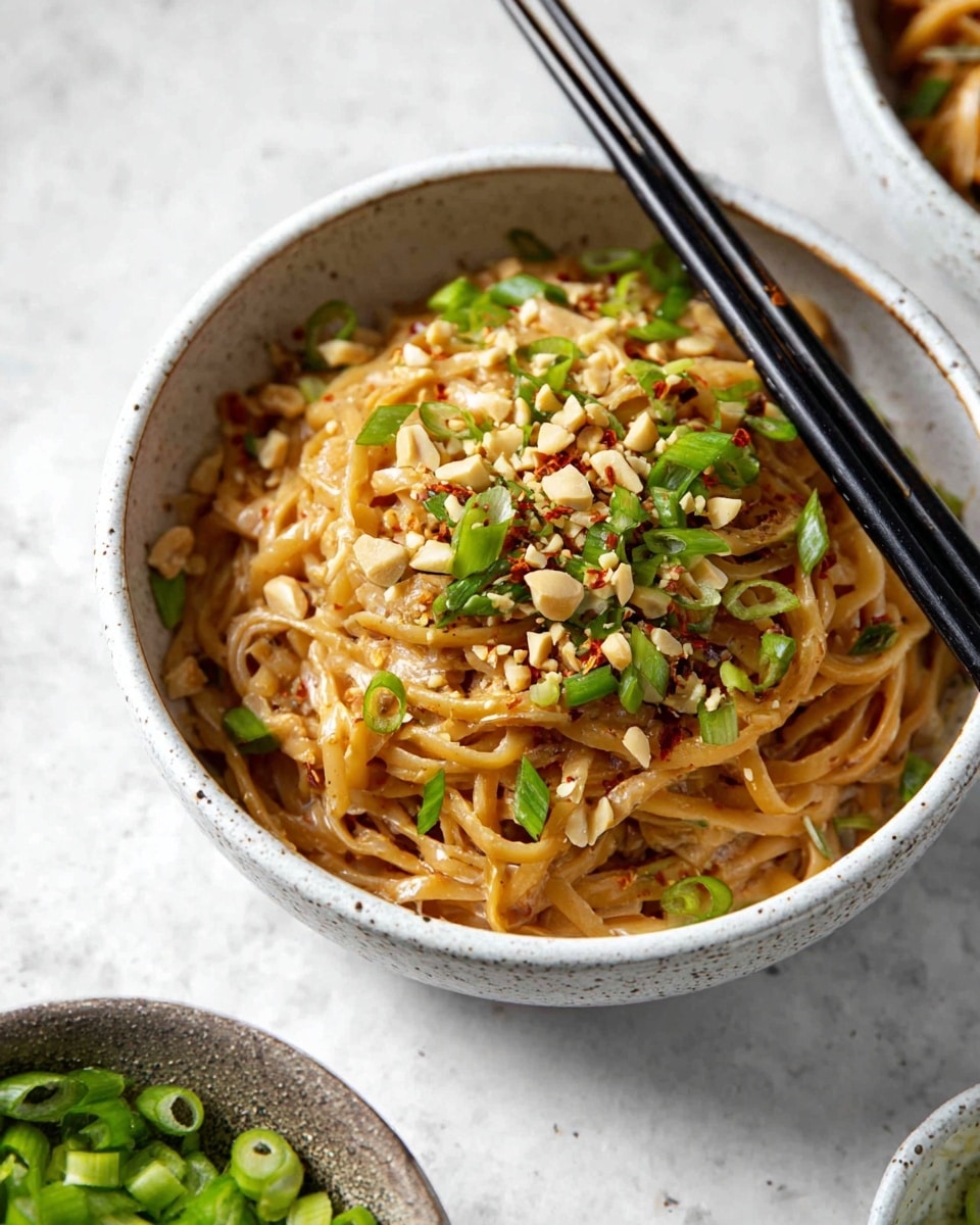 A bowl of thick, light brown noodles sits in a white speckled bowl, topped with scattered chopped peanuts and thin slices of green onions, adding green and creamy beige spots on top. Small red chili flakes are sprinkled over the dish, giving a hint of red color and texture contrast to the glossy noodles underneath. The bowl is placed on a white marbled surface, with black chopsticks resting beside it. In the background, there are small white bowls filled with green onions and chili flakes. photo taken with an iphone --ar 4:5 --v 7