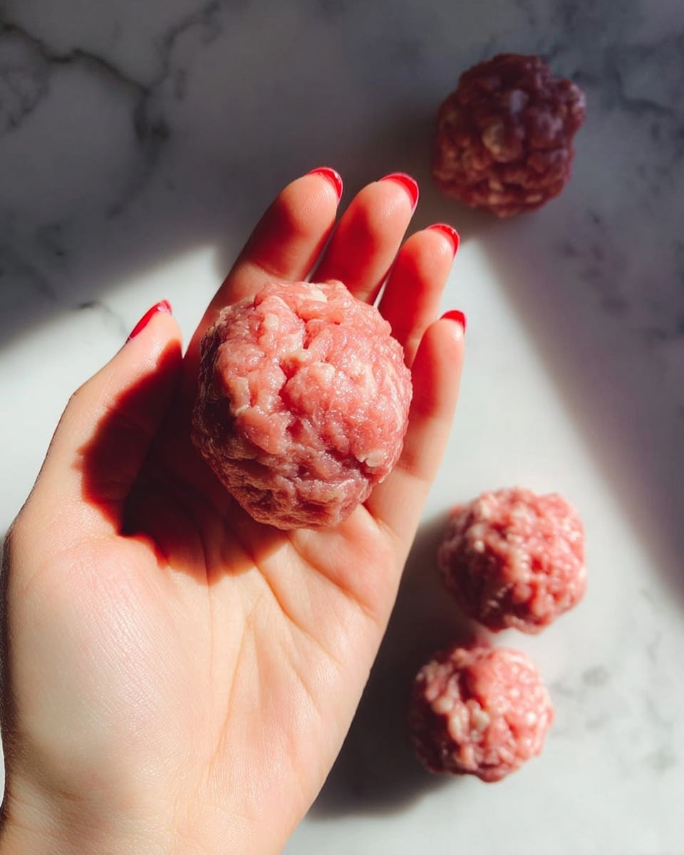 A close-up of a woman's hand holding a small round ball of raw ground meat showing its pinkish-red color with white fat bits mixed in. Below the hand, there is a white marbled surface where three more similar raw meat balls are loosely placed, casting soft shadows. The light creates a bright and natural look on the meat's texture and the woman's skin. photo taken with an iphone --ar 4:5 --v 7