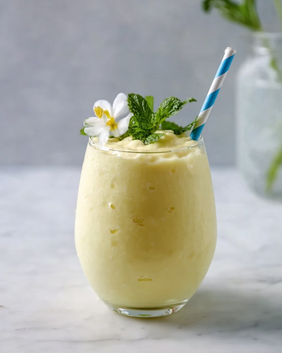 A creamy light yellow smoothie fills a clear rounded glass with a thick texture visible inside. On top, the smoothie is smooth with a small peak near the rim. A blue and white striped straw is placed upright in the middle of the drink. A small white flower with delicate petals and a tiny orange center rests on the foam next to a bright green mint leaf. The glass sits on a white marbled surface with soft natural light highlighting the soft colors and textures. photo taken with an iphone --ar 4:5 --v 7