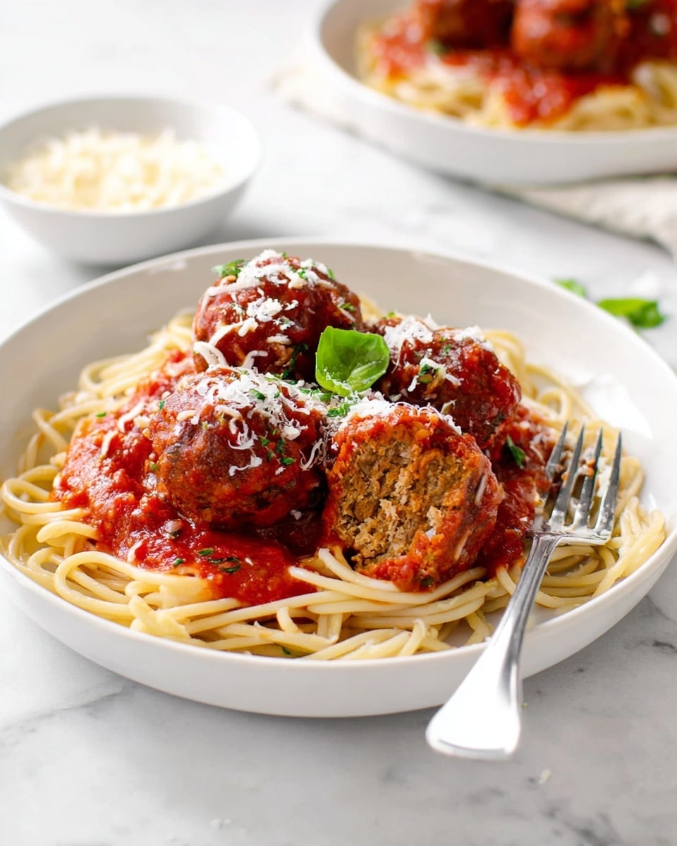 A white plate holds a neat round nest of plain light brown spaghetti at the bottom layer. On top, there are four large, round meatballs covered in thick bright red tomato sauce with visible texture. The meatballs are sprinkled with white shredded cheese and garnished with fresh green basil leaves at the center. A silver spoon and fork rest on the right side of the plate, slightly overlapping the spaghetti. The plate sits on a white marbled surface, and the background is softly blurred with hints of another similar plate and some green leaves. Photo taken with an iphone --ar 4:5 --v 7