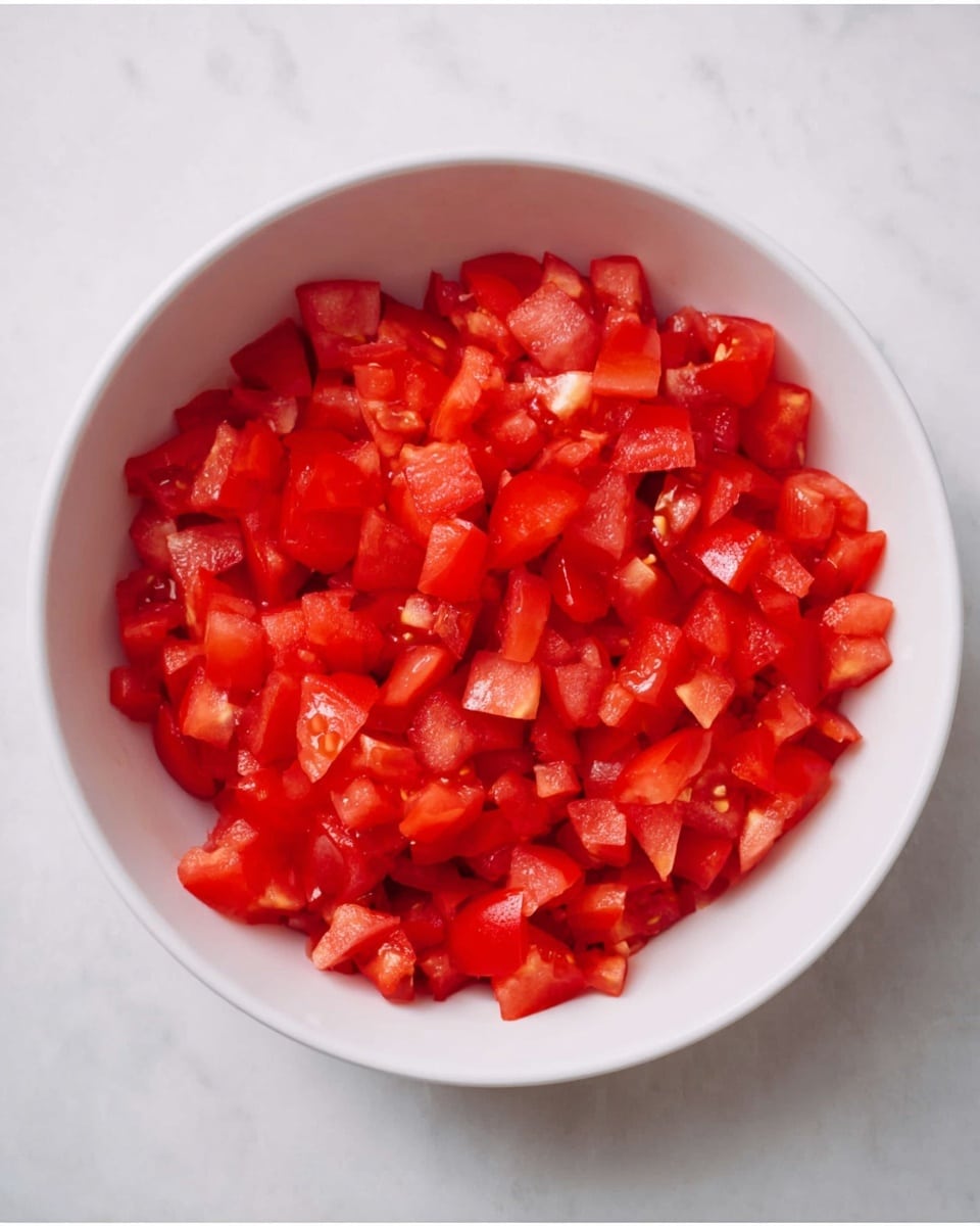 A white bowl filled with small, evenly diced bright red tomatoes, each piece showing a shiny and moist texture, evenly spread inside the bowl. The bowl is placed on a white marbled surface, creating a clean and simple look. photo taken with an iphone --ar 4:5 --v 7