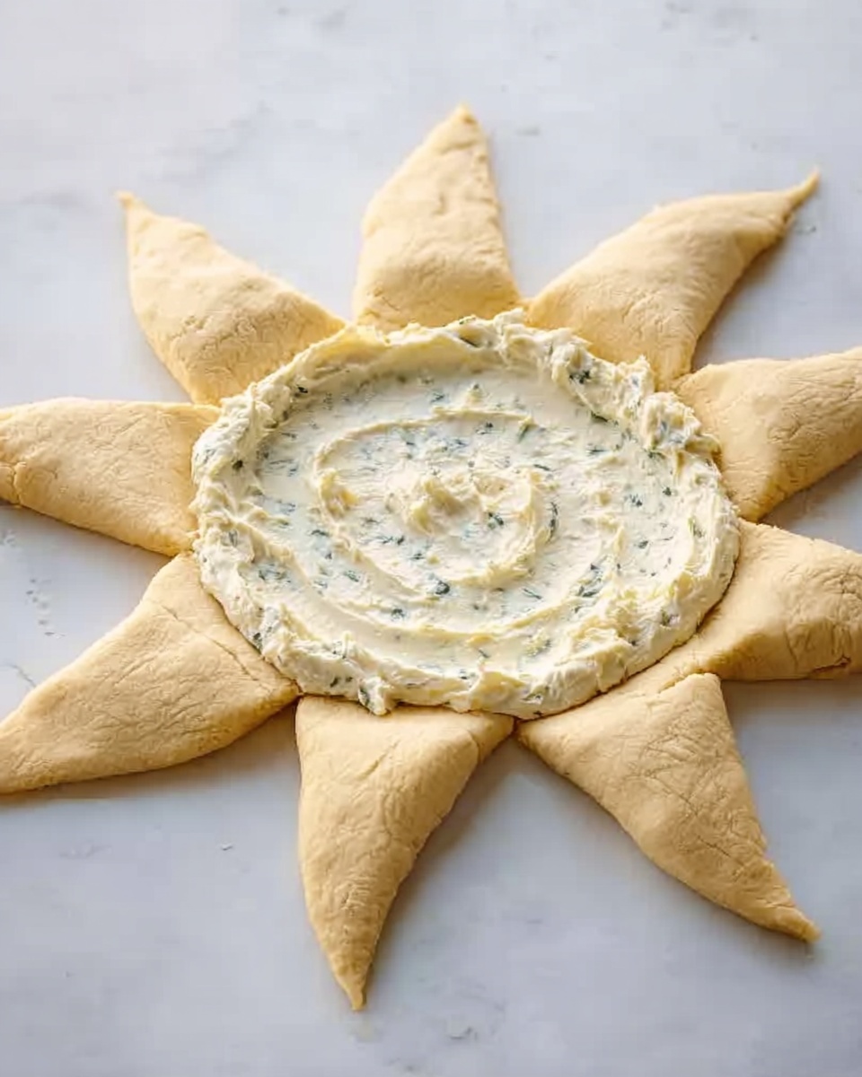 The image shows a sun-shaped dough on a white marbled surface. The dough is light golden and has eight pointed rays spreading outwards. In the center, there is a thick ring of white creamy spread with small green herb pieces mixed in, covering the middle area but leaving the rays uncovered. The texture of the dough looks soft and slightly dry, while the creamy spread appears smooth and thick. photo taken with an iphone --ar 4:5 --v 7