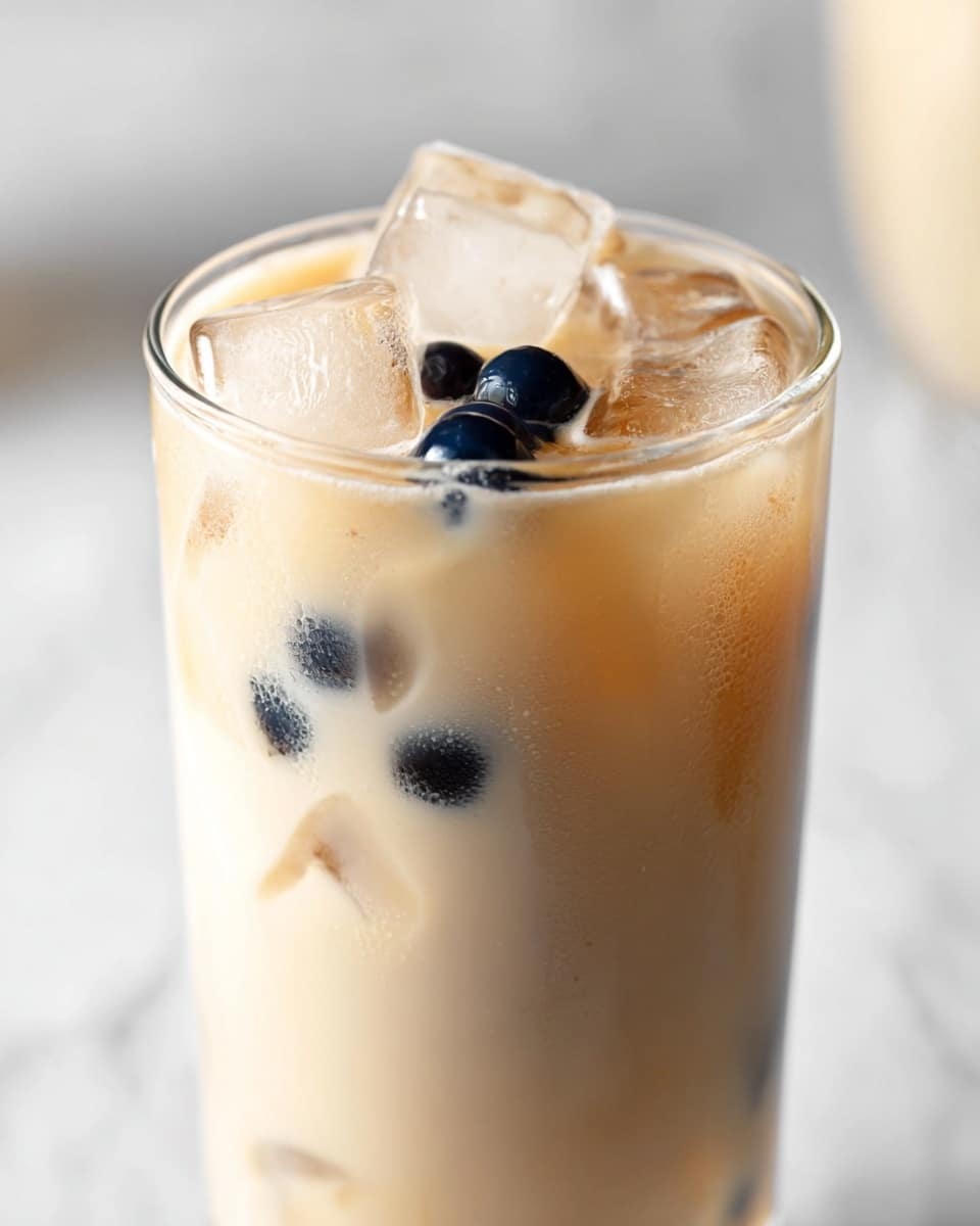 A tall, clear glass filled with a light beige creamy drink with a few black tapioca pearls floating near the top, mixed with several clear ice cubes that have slight cracks and bubbles inside. The drink has a smooth, milky texture visible beneath the ice, and a white marbled surface as the background. The focus is mainly on the top part of the glass showing the ice and pearls clearly, with the lower part of the drink slightly blurred. Photo taken with an iphone --ar 4:5 --v 7