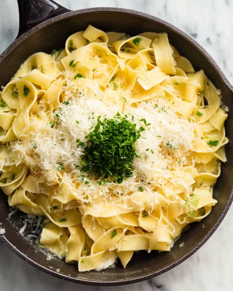 The image shows a close-up of a dark green cast iron pan filled with light yellow wide ribbon pasta. On top of the pasta, there is a pile of finely grated white cheese and a small bunch of bright green chopped herbs, both placed near the center. The pasta looks soft and shiny from some butter or sauce around the edges. The pan is resting on a white marbled surface. photo taken with an iphone --ar 4:5 --v 7