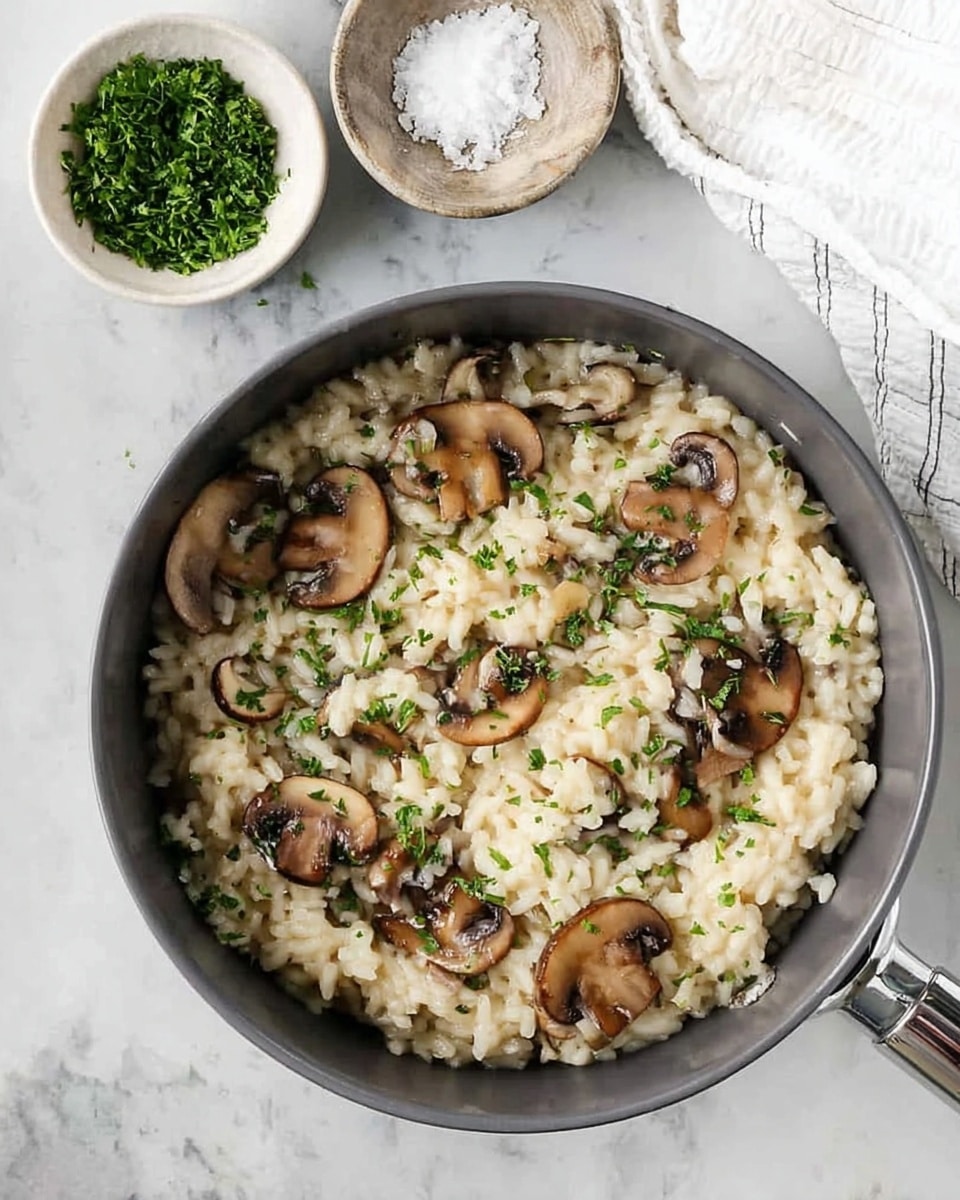 A dark gray pot filled with creamy mushroom risotto sits on a white marbled surface. The risotto is soft and pale cream in color, mixed with brown, sliced mushrooms topped with small bright green herb sprinkles, giving a fresh look. The texture of the rice is smooth and moist, with the mushrooms scattered evenly on top and within the rice. Next to the pot, two small white bowls hold chopped green herbs and coarse white salt, adding contrast to the scene. A white cloth napkin peeks in from the top right corner, with the shiny metal handle of the pot visible. Photo taken with an iphone --ar 4:5 --v 7