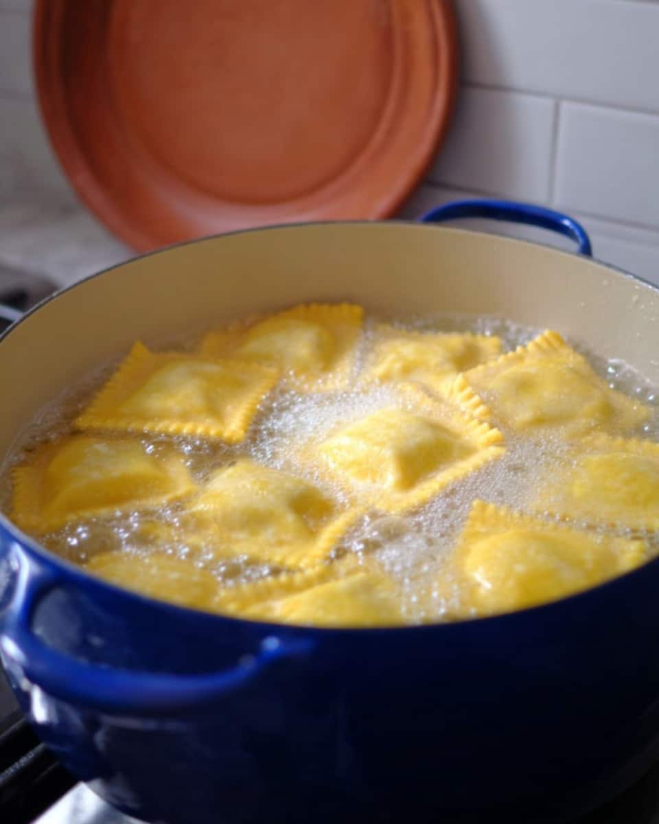 A blue pot filled with clear boiling water has around a dozen square yellow ravioli floating on top. The ravioli have a smooth texture with slightly crinkled edges and are submerged in bubbling water. In the background, a round terracotta lid rests against a white tiled wall, all set on a white marbled surface. photo taken with an iphone --ar 4:5 --v 7