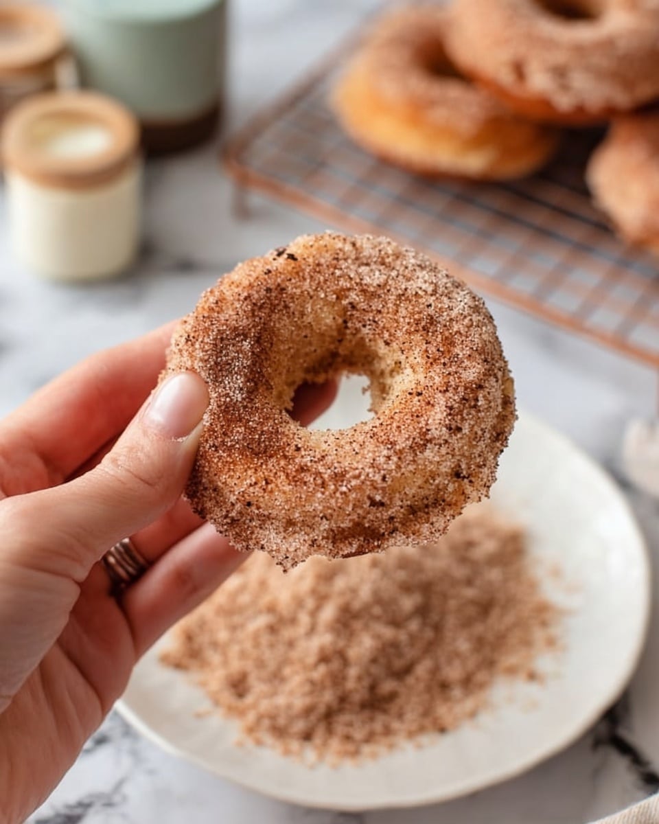 A close-up of a woman's hand holding a ring-shaped donut covered in a thick layer of cinnamon sugar, showing a rough, textured surface with some darker specks. Below, there is a white plate filled with more cinnamon sugar, slightly spread out in a circular shape with a soft, grainy look. The background features a white marbled surface with a blurred wire rack holding more donuts and small containers, creating a cozy baking atmosphere. Photo taken with an iphone --ar 4:5 --v 7