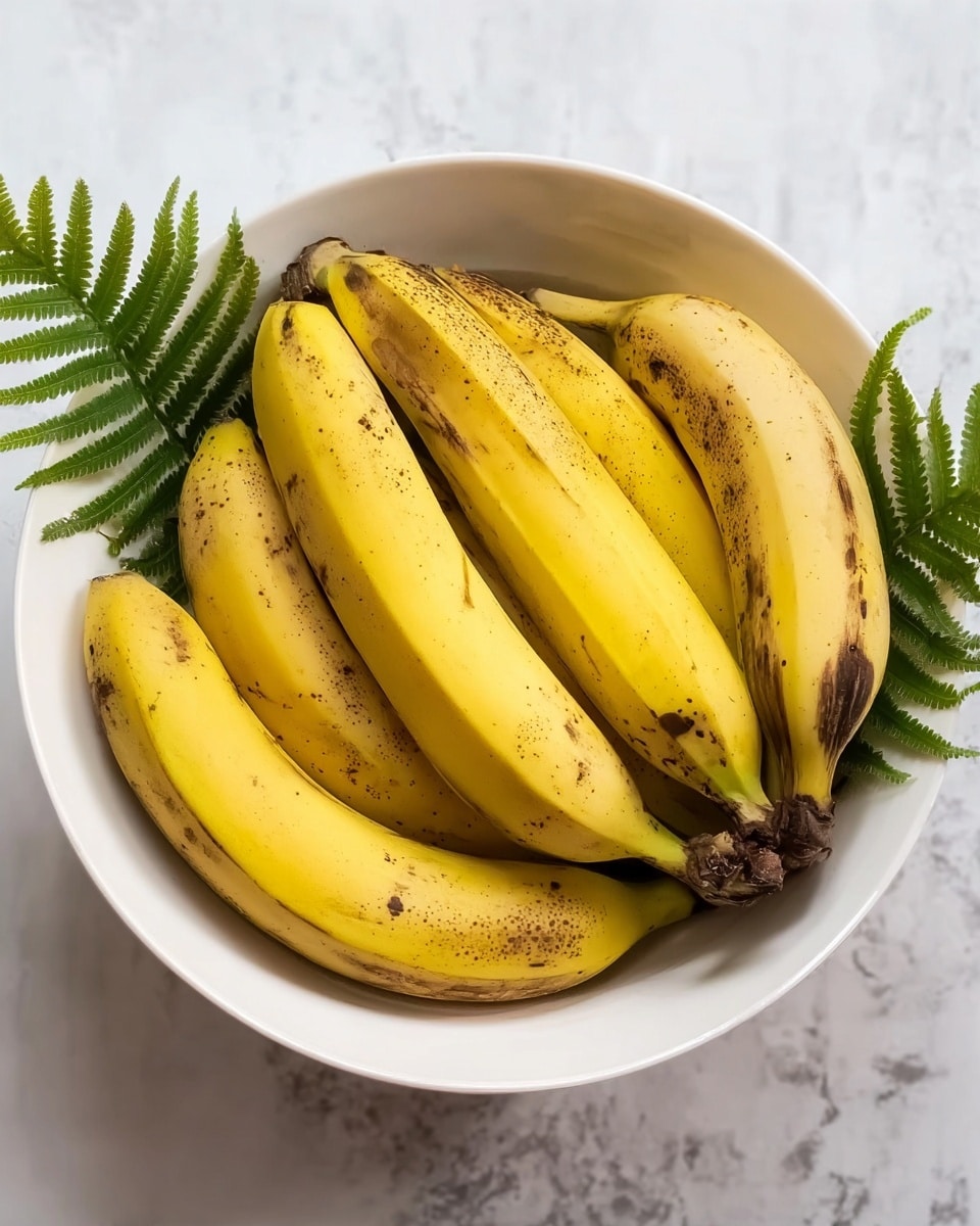 A white bowl filled with seven yellow bananas, each showing slight brown spots and marks, arranged so they curve upwards and lay close together, with some green fern leaves partly visible at the bottom inside the bowl. The bowl sits on a white marbled surface with soft lighting, creating a fresh and natural look. photo taken with an iphone --ar 4:5 --v 7