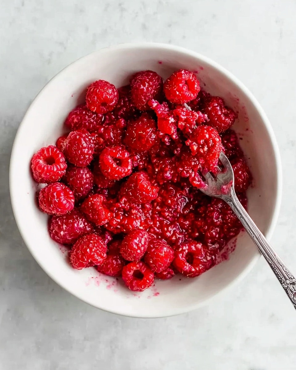A white bowl filled with vibrant red raspberries, some whole and some slightly mashed in the center, showing a mix of smooth and textured berry surfaces. A silver fork rests inside the bowl, touching the mashed raspberries, with its handle extending toward the bottom right of the bowl. The bowl is placed on a white marbled surface with faint veining, adding a subtle pattern to the background. photo taken with an iphone --ar 4:5 --v 7