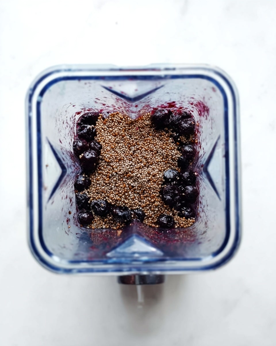 A clear blender container viewed from above shows a mixture with two main layers: the bottom layer contains dark purple whole berries, and the top layer is a generous covering of small, light brown seeds with a slightly rough texture. The container sits on a white marbled surface, highlighting the contrast between the dark berries and the grainy seeds. The blender's square shape with rounded edges is clearly visible. photo taken with an iphone --ar 4:5 --v 7