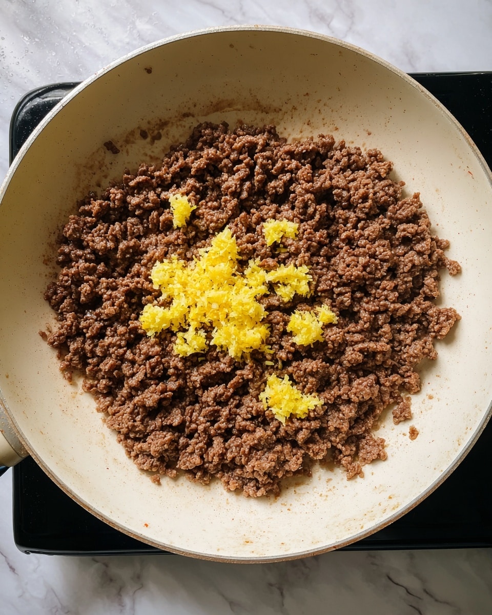 In a white pan, there is one layer of cooked brown ground meat with a slightly crumbly texture spread evenly across the surface. On top of this, in the center, are small piles of grated yellow ginger with a fibrous and moist texture, contrasting with the darker meat. The pan sits on a black stove surface, but the scene is set on a white marbled background. photo taken with an iphone --ar 4:5 --v 7