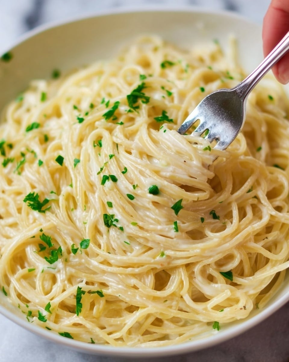 The image shows a close-up of creamy spaghetti pasta being mixed in a white bowl by a woman's hand holding a fork. The pasta is coated in smooth, light yellow sauce with a shiny texture. Small green parsley leaves are sprinkled evenly over the top, adding a fresh, colorful contrast. The noodles are long, thin, and intertwined in soft layers beneath the sauce. The bowl sits on a white marbled surface. Photo taken with an iphone --ar 4:5 --v 7