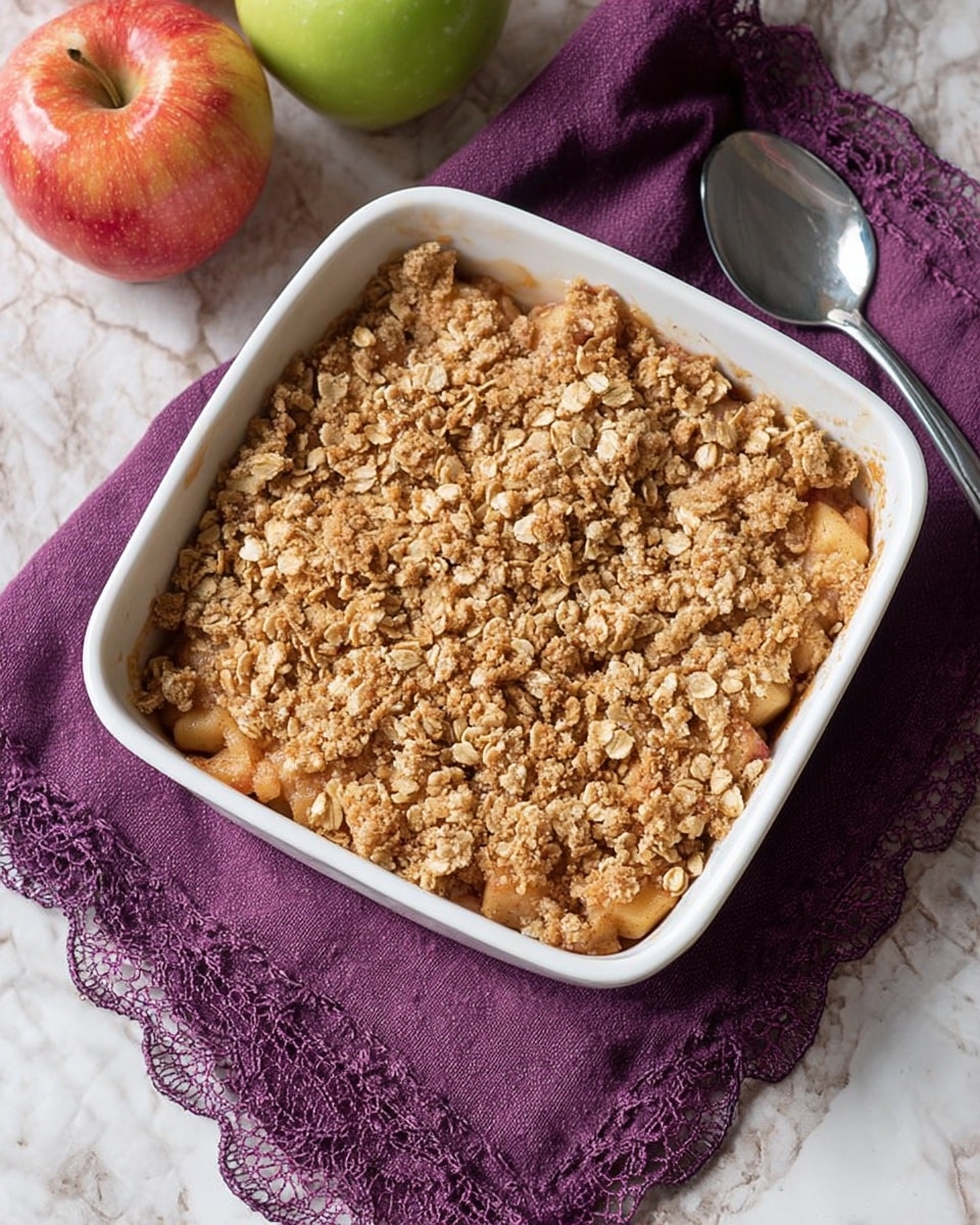 A white square baking dish filled with a two-layer dessert sits on a purple cloth with lace edges on a white marbled surface. The bottom layer is a chunky apple filling, showing soft pieces of reddish and greenish apple. The top layer is a golden-brown crumbly oat topping made of oats and sugar, scattered unevenly with some larger oat flakes. A silver spoon lies next to the dish, partly resting on the purple cloth. In the background, there are two whole apples, one red and yellow, and one green, placed close together. Photo taken with an iphone --ar 4:5 --v 7