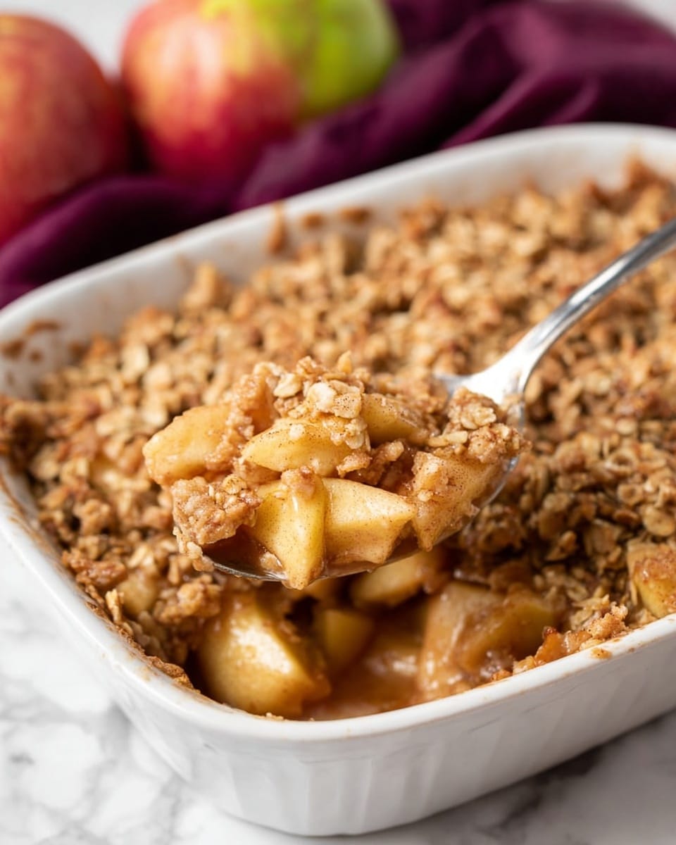 A white baking dish filled with a warm apple crisp, showing two layers: the bottom layer made of soft, chopped apple pieces mixed with cinnamon and juices, giving a light brown and slightly shiny look; the top layer is a crumbly oat topping with a rough, golden-brown texture. A silver spoon scoops into the dish, holding both apple pieces and oat topping. The background shows a slightly blurred purple cloth and some whole apples, all placed on a white marbled surface. photo taken with an iphone --ar 4:5 --v 7