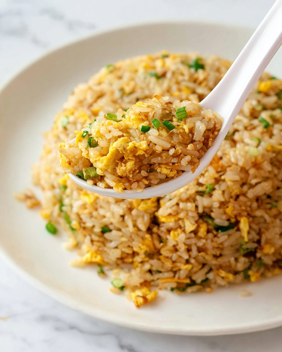 A white plate holds a large, round mound of fried rice made from long grains mixed with small pieces of scrambled yellow egg and bits of green onion. A white ceramic spoon lifts a spoonful of the rice close to the camera, showing the separate grains and the mix of yellow, light brown, and green colors with a slightly oily texture. The background is a white marbled surface, and the overall scene is brightly lit and focused on the spoonful of rice. Photo taken with an iphone --ar 4:5 --v 7