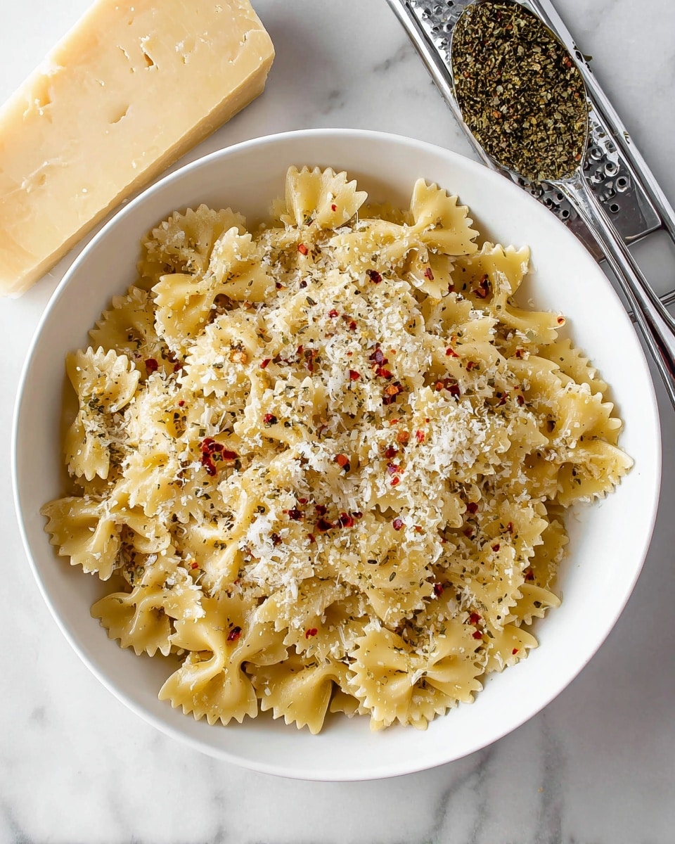 A white bowl filled with light yellow farfalle pasta layered evenly, sprinkled on top with finely grated white cheese and small bits of red chili flakes and black pepper, giving a speckled texture across the pasta shapes; the bowl sits on a white marbled surface with a block of pale yellow cheese and a metal grater on one side, and a spoonful of dried green herbs on the other side, adding visual interest around the bowl photo taken with an iphone --ar 4:5 --v 7
