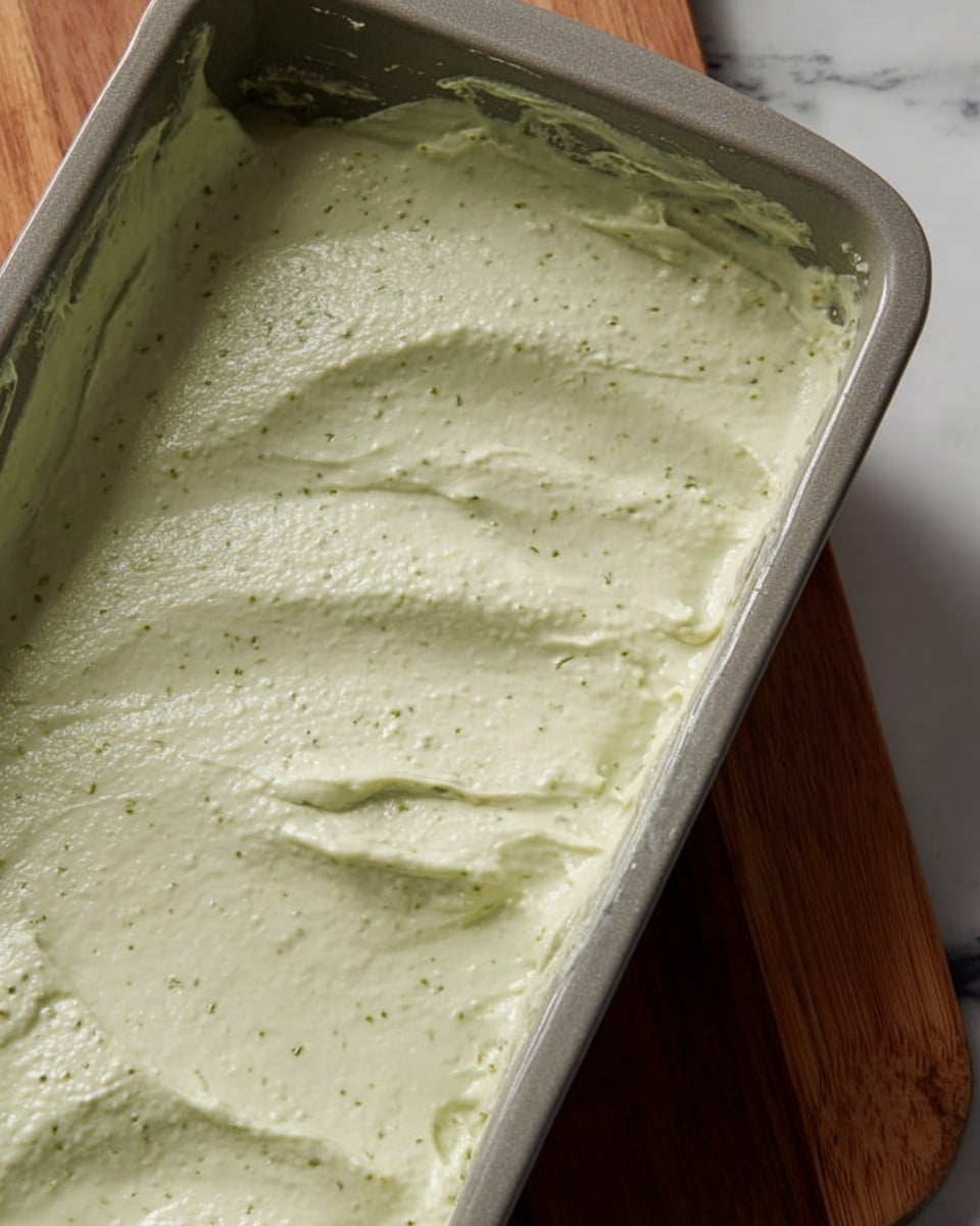 A close-up of a rectangular grey-green baking dish filled halfway with a thick, pale green creamy mixture that has small darker specks, spread unevenly with some texture bumps and smooth areas. The mixture is smeared along the bottom and partly up the sides of the dish, sitting on a white marbled surface with part of a wooden board beside it. photo taken with an iphone --ar 4:5 --v 7