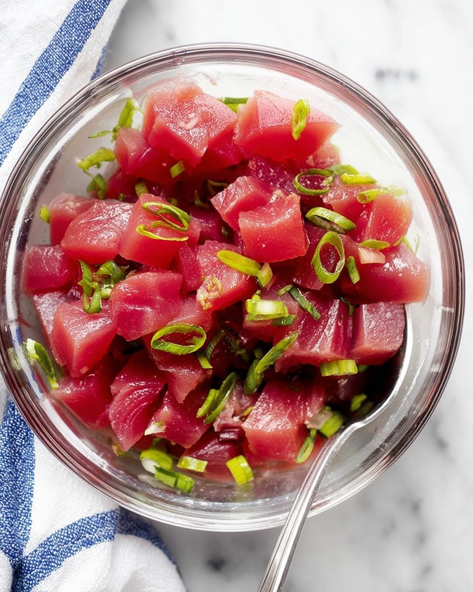 The image shows a clear glass bowl filled with small cubes of raw, deep pink tuna mixed with thin slices of bright green scallions. The tuna pieces have a smooth, fresh texture while the scallions add a fresh contrast with their slightly shiny, crisp look. A silver spoon is partially visible inside the bowl, resting among the tuna and scallions. The bowl sits on a white marbled surface with a portion of a blue and white striped cloth napkin visible beside it. photo taken with an iphone --ar 4:5 --v 7