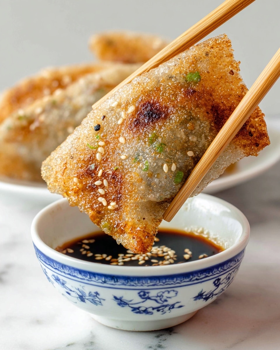 A close-up of a golden-brown, crispy fried dumpling covered with white sesame seeds and small green onion pieces. The dumpling's texture is bubbly and slightly shiny, showing a mix of soft and crunchy parts, with darker brown spots from frying. It is held by a pair of wooden chopsticks and is being dipped into a small white bowl with blue decoration, filled with dark soy sauce. The background is a clean white marbled surface. A woman's hand holds the chopsticks gently, with the dumpling angled toward the sauce. photo taken with an iphone --ar 4:5 --v 7