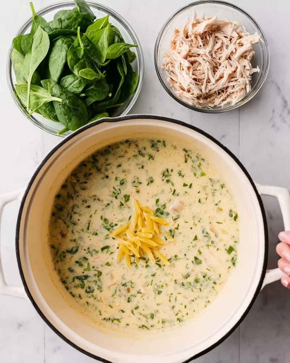 The image shows a white pot with black trim filled with creamy soup. The soup has small green herb pieces mixed throughout and a few yellow pasta pieces floating near the top. Next to the pot, there are two small clear bowls placed on a white marbled surface. One bowl contains fresh green spinach leaves, and the other has shredded chicken. A woman's hand is arranging the items, contributing to a lively cooking scene. Photo taken with an iphone --ar 4:5 --v 7