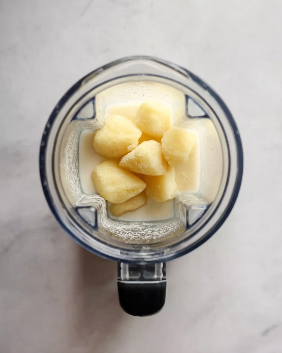 Top view of a clear blender filled with two layers: the bottom layer is a milky white liquid with a smooth texture, and the top layer consists of several pale yellow frozen fruit chunks with a slightly rough surface. The blender is placed on a white marbled surface, giving a clean and bright look. The blender container has clear sides and a black handle. Photo taken with an iphone --ar 4:5 --v 7