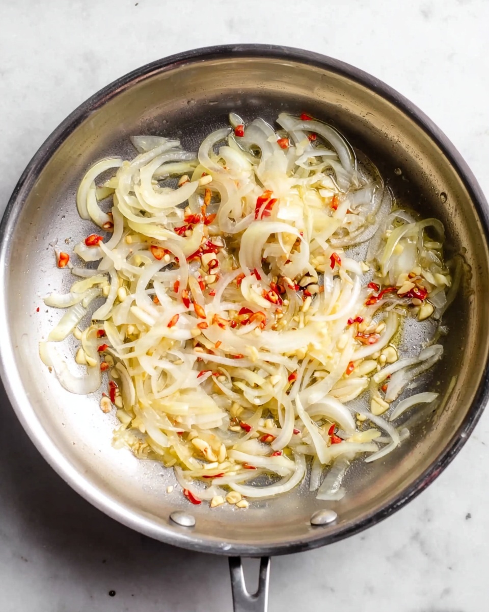 The image shows a top view of a metal frying pan placed on a white marbled surface. Inside the pan, there are thin slices of light yellow onions spread across the base, mixed with small bits of red chili and thin slices of garlic scattered evenly throughout. The onions are slightly glossy, indicating they are being lightly cooked, and the red chili bits add a splash of vibrant color. The pan has a reflective surface that contrasts with the soft textures of the cooking ingredients. Photo taken with an iphone --ar 4:5 --v 7