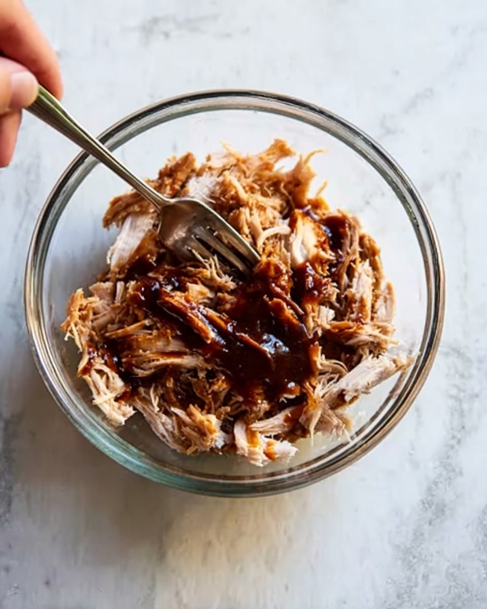 A clear glass bowl holds shredded cooked meat in the center, with a dark brown sauce drizzled over the top, creating a glossy texture. A fork is placed inside the bowl on the right side, appearing to be ready to mix or pick up the food. The bowl rests on a white marbled surface, and part of a woman's hand can be seen in the top left corner, lightly touching the bowl. The lighting is soft and natural, giving the dish a warm and inviting appearance. photo taken with an iphone --ar 4:5 --v 7