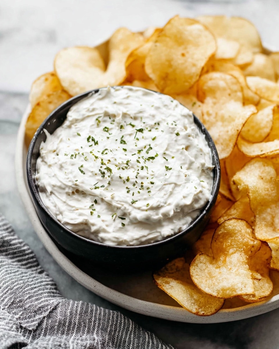 A close-up view shows a dark bowl filled with thick white dip, topped with small green herb sprinkles scattered across the surface. The bowl sits on a larger white bowl that holds many light golden, crispy potato chips, which have curved and slightly wrinkled textures. The background is a white marbled surface with a soft striped gray and white cloth placed nearby. The photo taken with an iphone --ar 4:5 --v 7