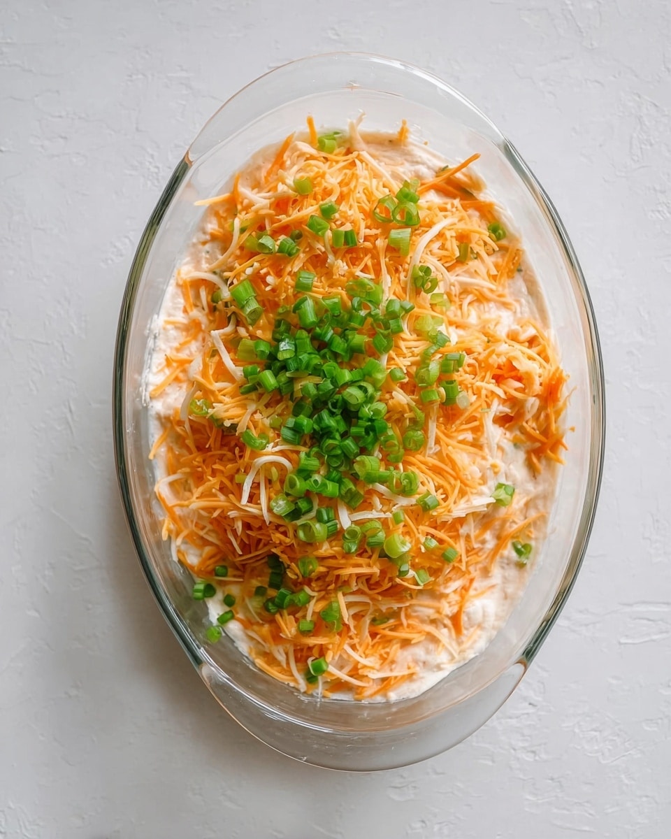 A clear glass oval dish holds a two-layer mixture, starting with a creamy pale base visible at the edges, topped with a generous layer of shredded cheese that shows orange and white strands mixed together. On top of the cheese, there is a scattered small pile of bright green chopped scallions, adding a fresh contrast. The dish sits on a flat surface with a white marbled texture, and the entire scene is viewed from above. Photo taken with an iphone --ar 4:5 --v 7