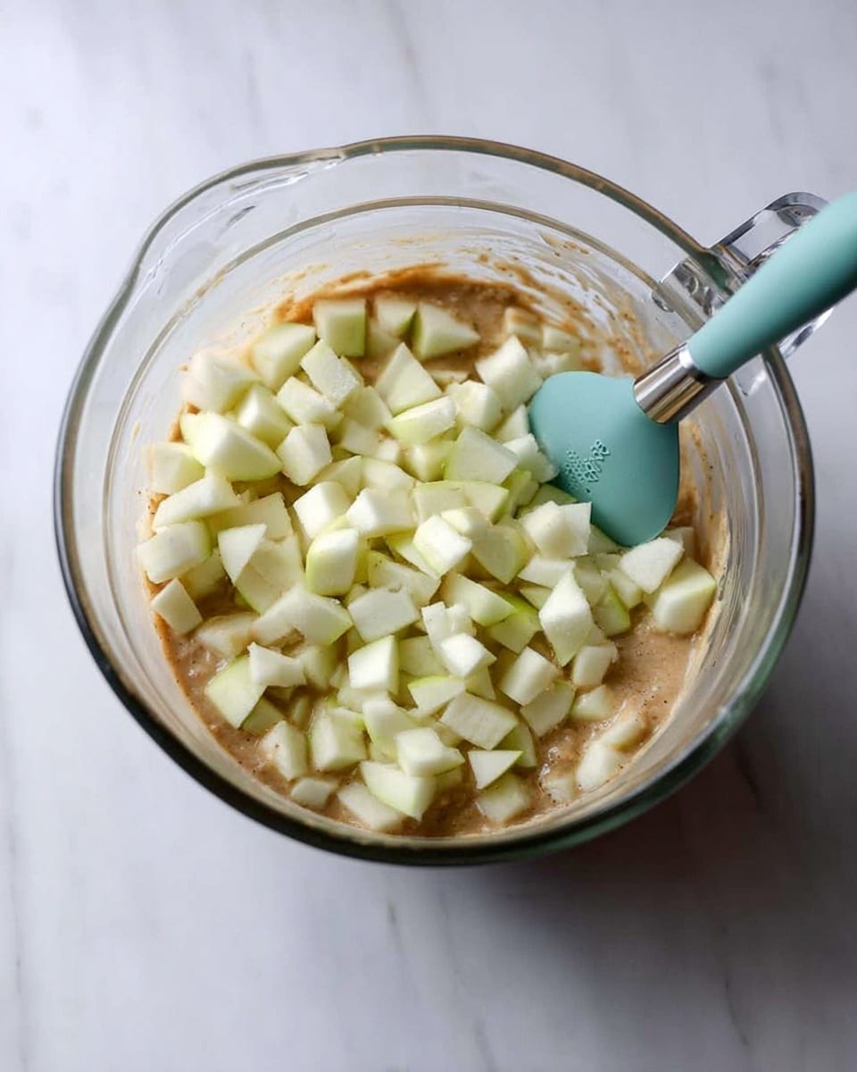A clear glass measuring cup filled with light brown batter as the bottom layer, topped with many small white and pale green apple chunks scattered evenly across the surface. A teal silicone spatula with a silver handle is resting inside the cup, touching the batter and apple pieces. The cup sits on a white marbled surface. photo taken with an iphone --ar 4:5 --v 7