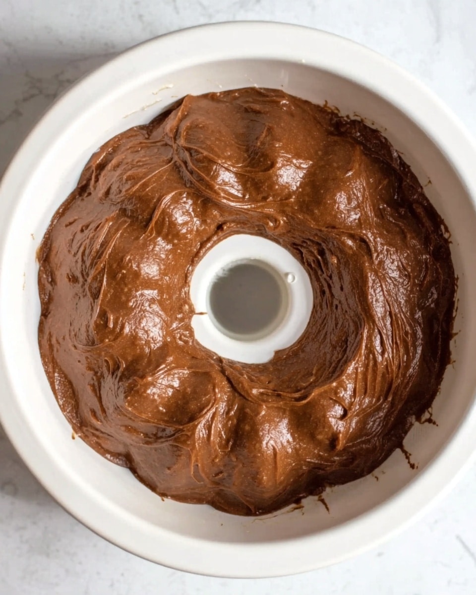 The image shows a white bundt cake pan filled with a thick, smooth chocolate batter spread evenly inside. The batter is dark brown in color with a shiny, wet texture and fills the pan to the edges but does not overflow. The pan has a central hole creating a ring shape for the batter. The pan sits on a white marbled surface. Photo taken with an iphone --ar 4:5 --v 7