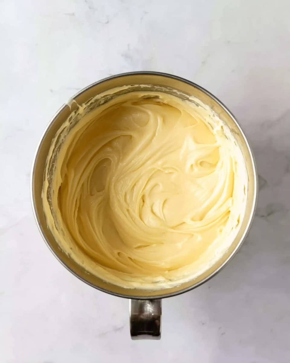 A top view of a silver metal mixing bowl filled with creamy, smooth, pale yellow batter with soft swirls on the surface. The bowl is centered on a white marbled textured surface. The metal bowl has a small loop handle at the top and a short spout at the bottom edge. photo taken with an iphone --ar 4:5 --v 7