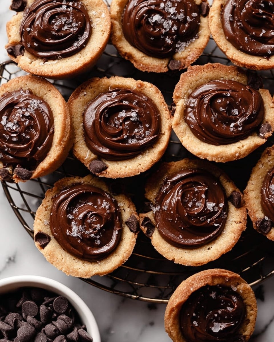 The image shows a group of small tart-like cookies arranged closely on a dark wire rack over a white marbled surface. Each cookie has two visible layers: the bottom layer is a light brown, thick, slightly uneven cookie crust with chocolate chips embedded in it, and the top layer is a smooth, glossy, dark chocolate filling spread in a swirl pattern. Some cookies appear sprinkled lightly with coarse salt crystals on the chocolate. In the bottom left corner, a white bowl filled with small dark chocolate chips is partially visible. The overall composition highlights the contrast between the rough cookie edge and shiny chocolate center, with soft light enhancing the textures. Photo taken with an iphone --ar 4:5 --v 7
