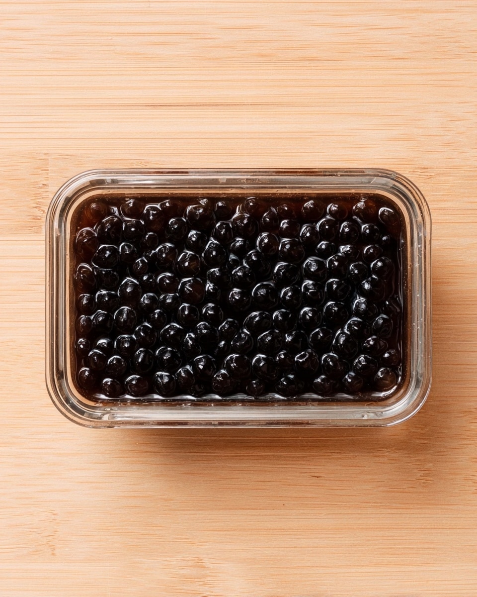The image shows a small rectangular clear glass container filled with shiny, black tapioca pearls in a dark syrup liquid. The pearls are packed closely together, creating a uniform layer with a glossy texture. The container is placed on a light-colored wood surface, visible around all sides of the container. photo taken with an iphone --ar 4:5 --v 7