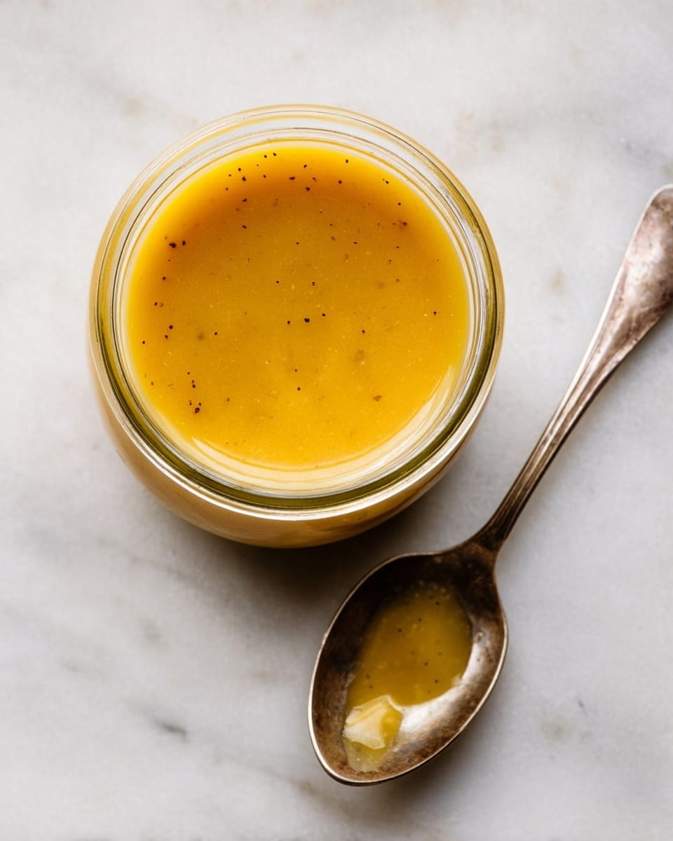 A clear glass jar filled with a smooth, bright golden-yellow sauce with tiny black specks floating inside, placed on a white marbled surface. Next to the jar, a tarnished silver spoon lies flat with a small amount of the same golden sauce coating its bowl, showing a glossy texture. The photo is taken from above, focusing on the open jar's round mouth and the shiny spoon close by, with soft natural light highlighting the sauce's rich color photo taken with an iphone --ar 4:5 --v 7