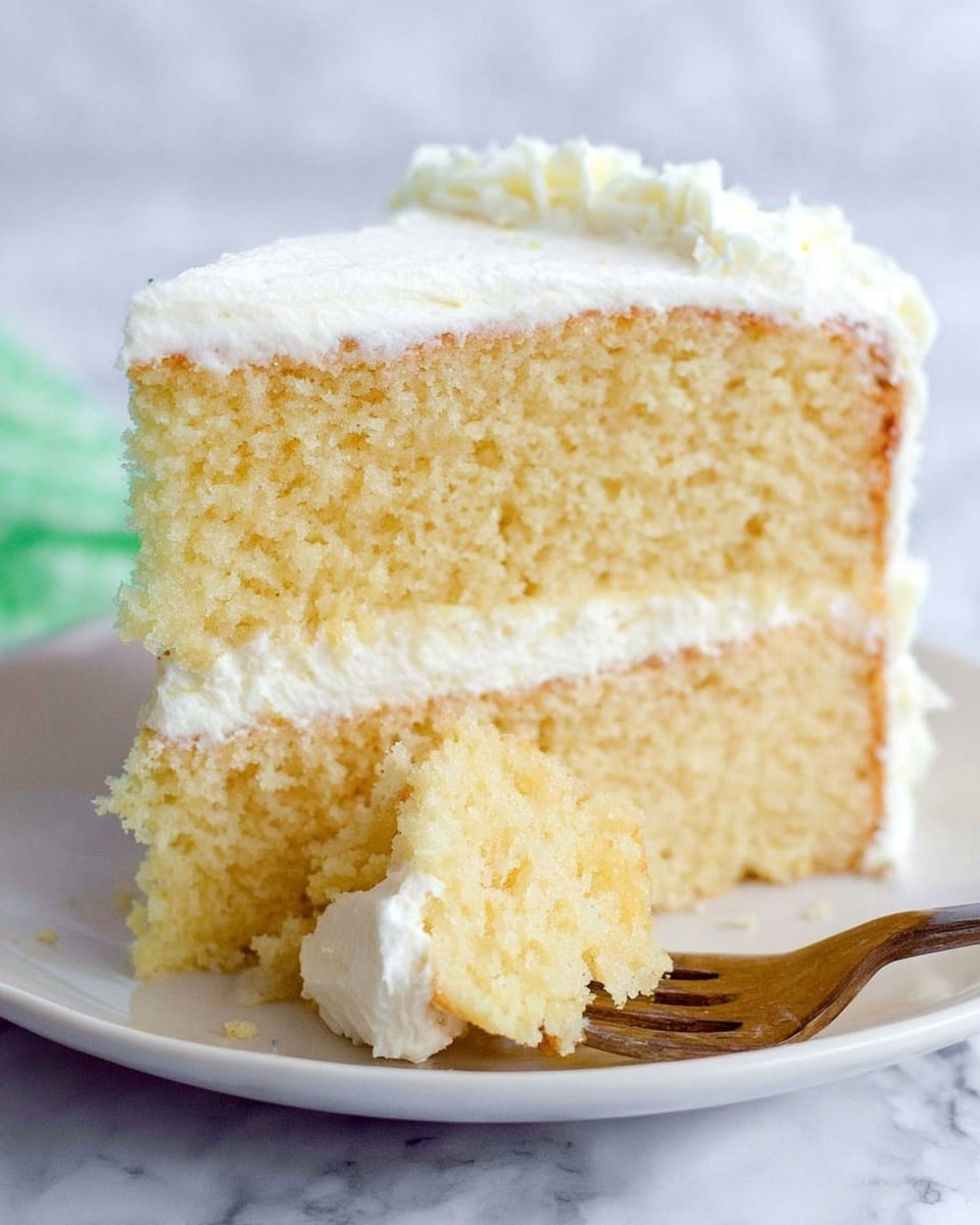 A slice of two-layer yellow cake with a light, fluffy texture is shown on a white plate. Between the layers and on top is a thick layer of smooth white frosting, slightly whipped. The fork resting on the plate holds a bite-sized piece of the cake with frosting. The background is a white marbled texture. photo taken with an iphone --ar 4:5 --v 7