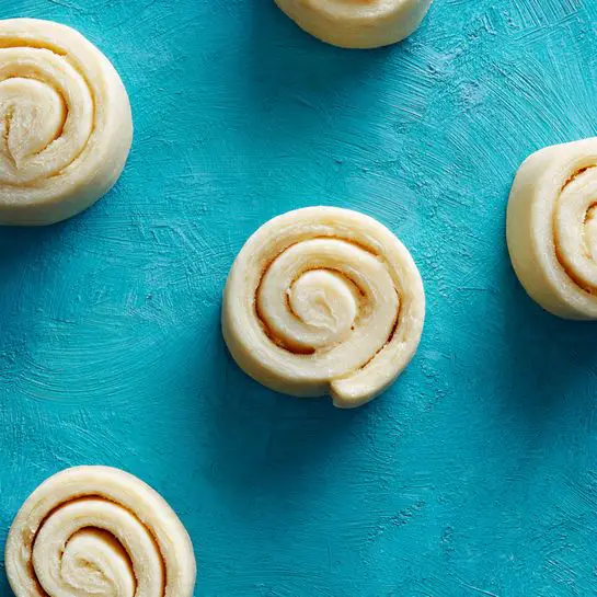 The image shows several small rolled dough pieces placed on a textured blue surface, with one row partially aligned and two separate pieces placed closer to the camera. The dough appears light beige and slightly soft with visible swirled layers inside each small roll, indicating a spiral shape. The surface is smooth but has a subtle pattern, adding depth to the image. The overall look is raw and ready to be cooked or baked. Photo taken with an iphone --ar 4:5 --v 7