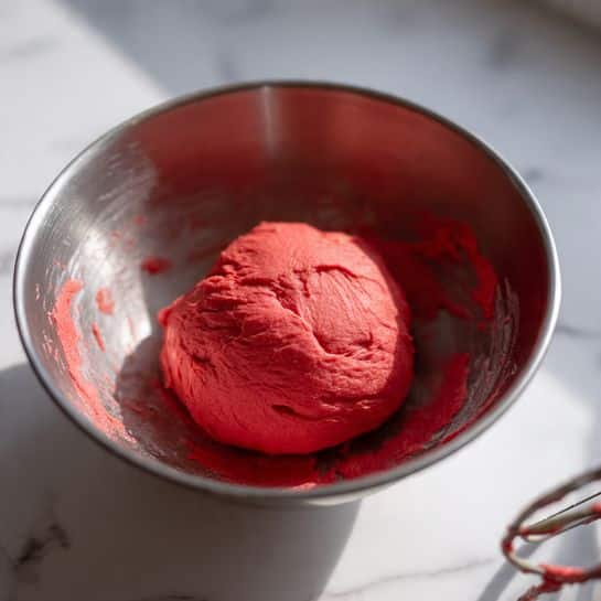 A close-up view of a single, smooth ball of bright red dough sitting inside a clear metal mixing bowl with some dough residue on the sides. The dough has a soft, slightly shiny texture with small folds and creases, making it look freshly mixed and elastic. The bowl is placed on a white marbled surface, and part of a mixing tool is visible blurred in the front right corner. photo taken with an iphone --ar 4:5 --v 7