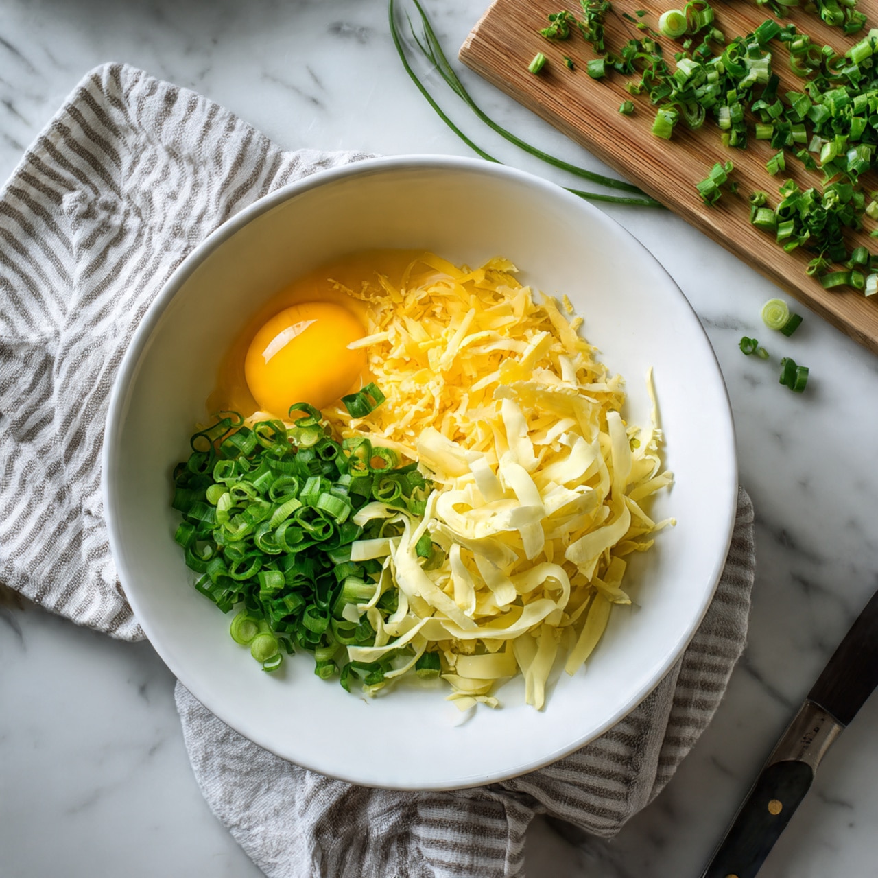 A white bowl sits on a white marbled surface with a striped cloth underneath it. Inside the bowl, there are three raw egg yolks positioned together on the lower left side, shredded pale yellow cheese piled high on the right side, and chopped green onions placed on the top left side. Above the bowl, to the right, there is a wooden cutting board with more chopped green onions and a knife with a wooden handle resting on it. A fork lies on the right side of the bowl on the surface. The scene is bright and clean. photo taken with an iphone --ar 4:5 --v 7