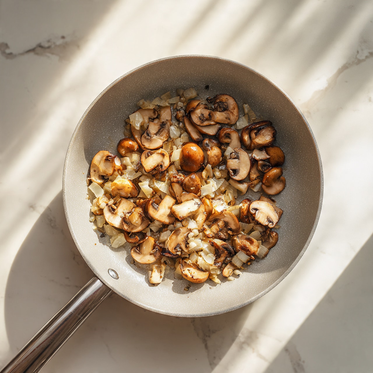 A top view of a light gray frying pan with a metal handle filled with sautéed mushrooms and finely chopped onions. The mushrooms are sliced into halves and quarters, showing their brown and beige colors with a slightly crispy texture on the edges. The chopped onions are golden brown and mixed evenly with the mushrooms, scattered across the pan's bottom. The pan rests on a white marbled surface with warm sunlight casting soft shadows around it. photo taken with an iphone --ar 4:5 --v 7
