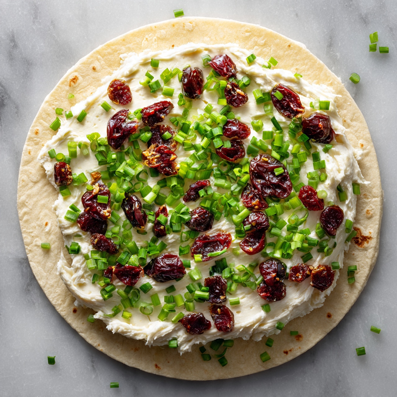 A round flatbread is spread with a smooth, white cream layer covering the entire surface. On top of the cream, small chopped green onions are scattered evenly, adding bright green spots all over. Mixed in with the green onions are small, dark red dried berries, arranged randomly across the flatbread. The flatbread is set on a white marbled surface with some dried berries blurry in the background. photo taken with an iphone --ar 4:5 --v 7