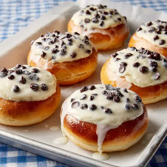 Five round pastries are shown on white parchment paper over a metal pan with a blue and white checkered cloth below. Each pastry has a golden brown, soft, and slightly puffy base. On top of each base is a thick creamy white layer that looks smooth and slightly melted. Scattered unevenly across this creamy layer are small, shiny dark chocolate chips. A white glaze is lightly drizzled over the pastries, creating small drips down the sides and spots on the parchment paper. The overall arrangement is casual with the pastries close to each other but not touching. photo taken with an iphone --ar 4:5 --v 7