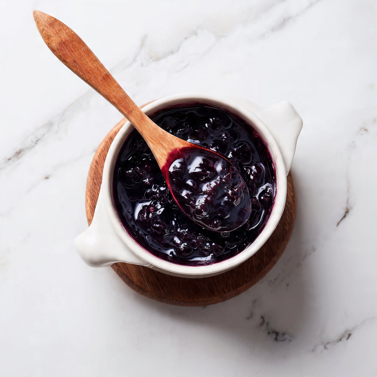 A white pot with a wooden handle holds a shiny dark purple sauce with small whole berries floating in it. A wooden spoon lifts some thick sauce, showing its smooth, glossy texture with deep purple and red shades. The inside edge of the pot has streaks of sauce. The pot sits on a small round trivet on a white marbled surface. Photo taken with an iphone --ar 4:5 --v 7