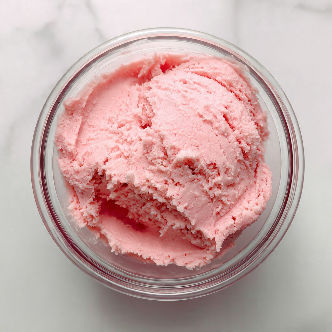 The image shows a close-up of soft, light pink dough with a slightly fluffy texture. The dough looks smooth but has some small ridges and uneven parts from being mixed. The dough fills most of a clear glass container, and the background is a white marbled surface. The lighting is bright, making the pink color stand out clearly. The focus is mainly on the center of the dough, showing its soft and creamy look photo taken with an iphone --ar 4:5 --v 7