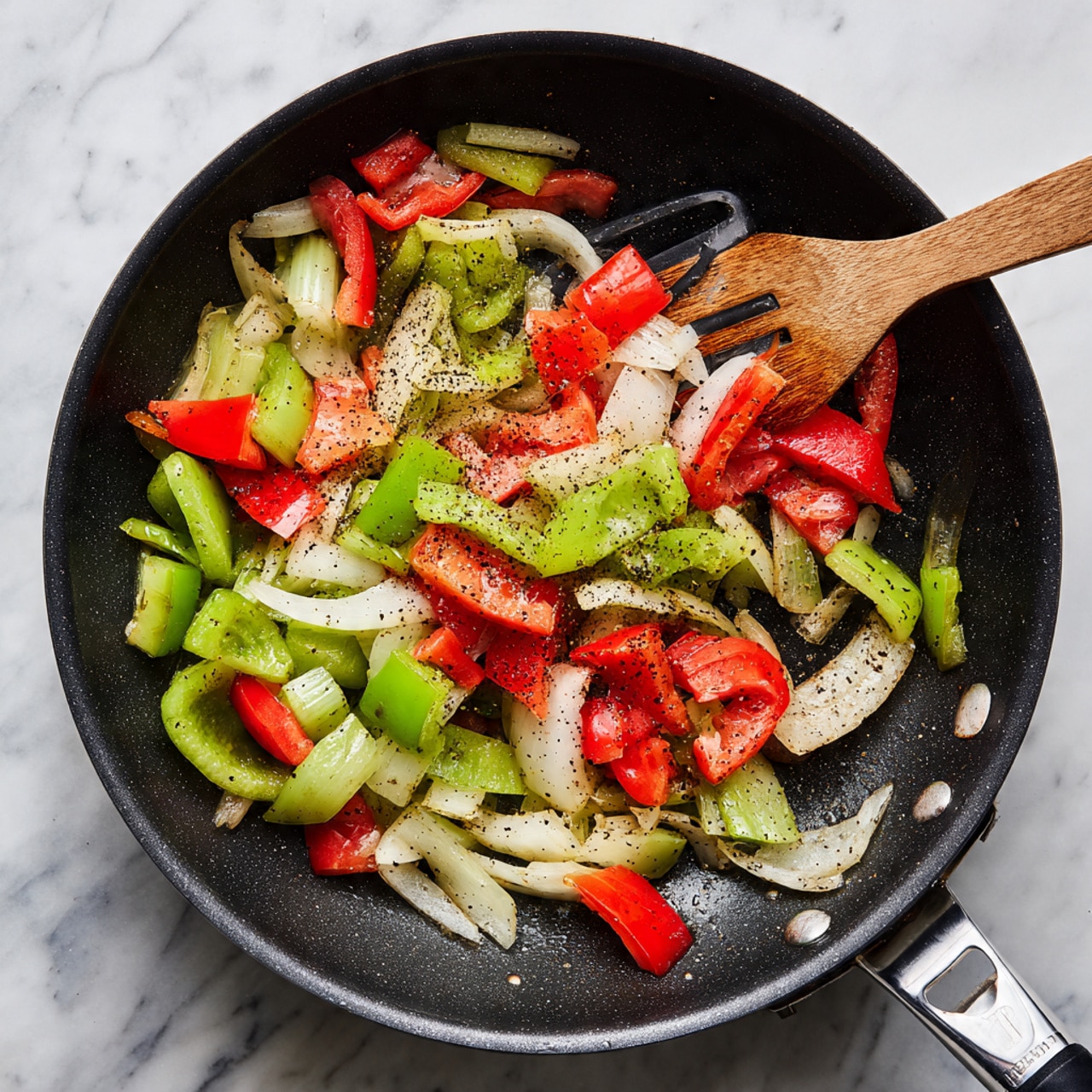 Inside a black pan, there is one layer of cooked vegetables including white onion slices, bright red bell pepper chunks, and light green celery pieces, all mixed with black pepper and small bits of seasoning. A metal spatula is partly visible in the pan, stirring the vegetables. The background and surface have a white marbled texture. Photo taken with an iphone --ar 4:5 --v 7
