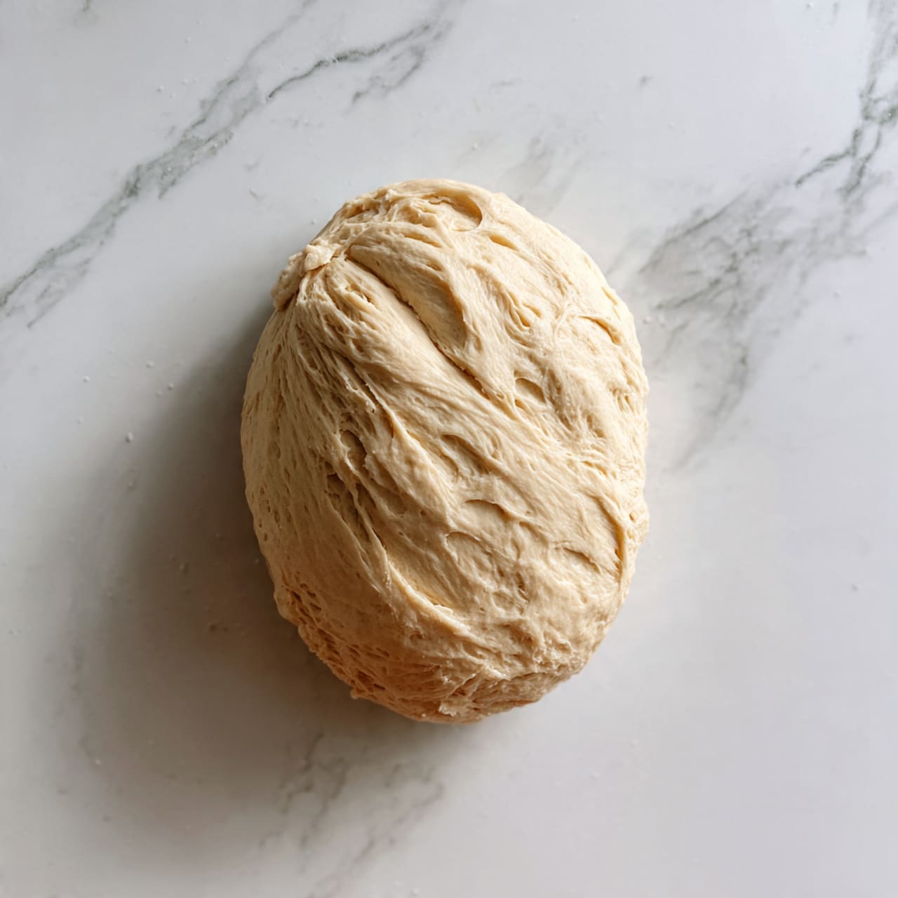 The image shows an uncooked piece of dough placed on a white marbled surface. The dough is oval-shaped with a smooth, slightly uneven texture and pale beige color. The surface of the dough has small dimples and slight indentations, indicating it has been stretched or pressed by a woman's hand. The dough looks soft and elastic, ready for baking or further preparation. Photo taken with an iphone --ar 4:5 --v 7