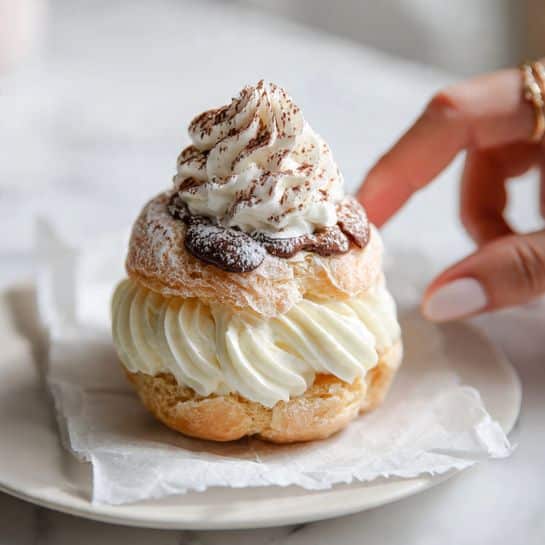 The image shows a cream puff dessert with three clear layers: the bottom layer is a light golden puff pastry with a soft flaky texture, the middle layer is thick white whipped cream swirled evenly, and the top layer is another puff pastry covered with powdered sugar and a small swirl of whipped cream dusted lightly with cocoa powder. The dessert is placed on parchment paper inside a white plate with rounded edges, set on a white marbled surface. A woman's hand is reaching toward the cream puff from the right side. Photo taken with an iphone --ar 4:5 --v 7