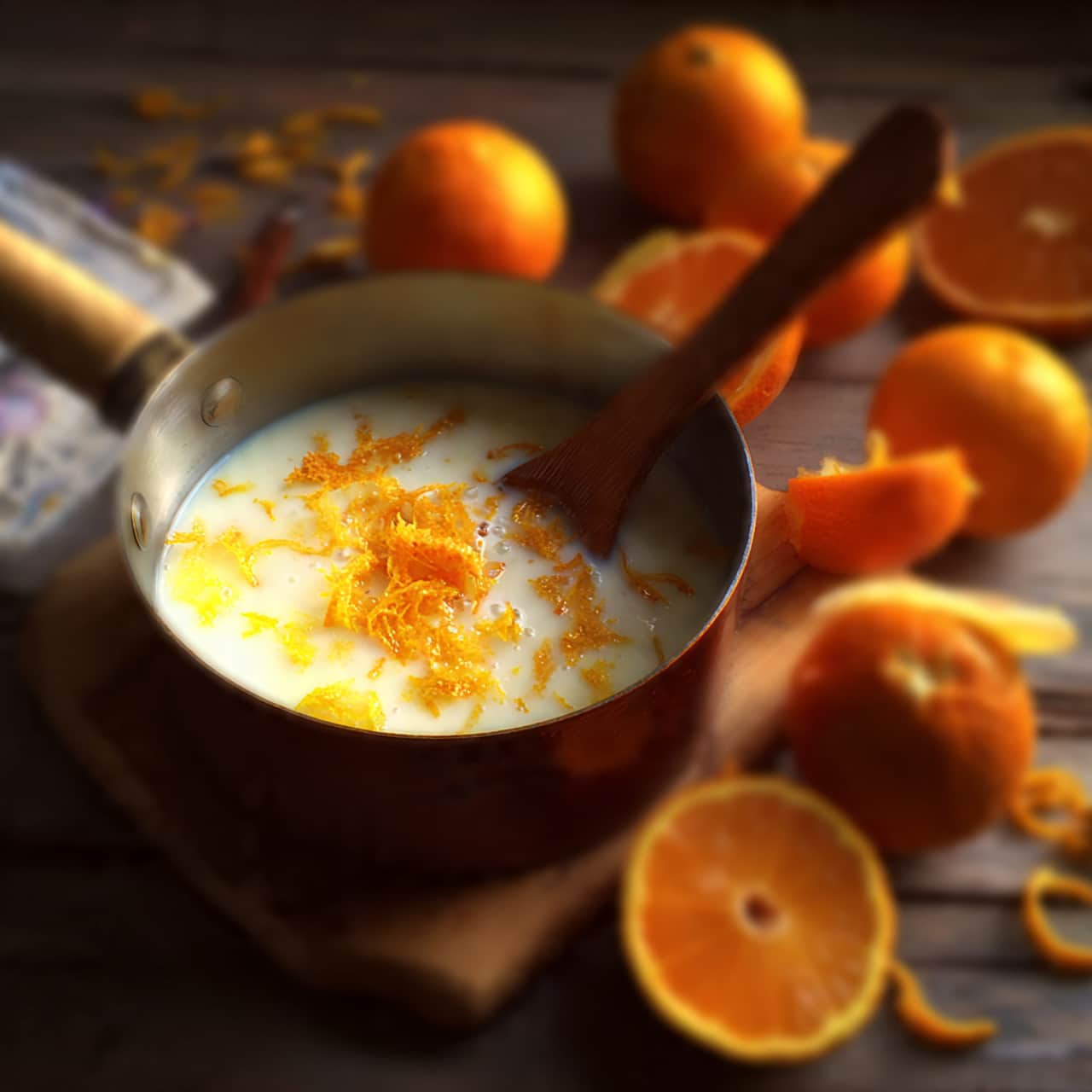 A close-up image shows a small silver pot filled with white milk. Inside the milk, there are a few orange peel pieces floating. A woman's hand with red nail polish is stirring the milk gently using a small white spatula with a wooden handle. The pot rests on a white marbled surface, and there are whole and half-cut oranges visible around the pot. The lighting is bright and natural, casting soft shadows. photo taken with an iphone --ar 4:5 --v 7